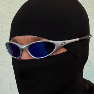 May include: Silver-framed sunglasses with blue lenses are worn over a black balaclava. The sunglasses have a sleek, modern design. The balaclava covers the head and neck, leaving only the eyes visible.