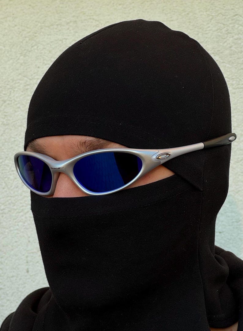 May include: Silver-framed sunglasses with blue lenses are worn over a black balaclava. The sunglasses have a sleek, modern design. The balaclava covers the head and neck, leaving only the eyes visible.