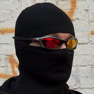 Op de afbeelding: Zwarte bivakmuts en zonnebril met rood en geel gespiegelde glazen. De zonnebril heeft een zwart montuur met een rood logo. De achtergrond is een verweerde bakstenen muur.