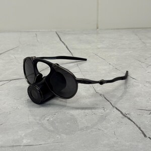 May include: Black sunglasses with a unique design. The sunglasses feature round lenses and a cylindrical component between the lenses. The arms of the sunglasses are thin and curve around the ears. The sunglasses are resting on a gray surface.
