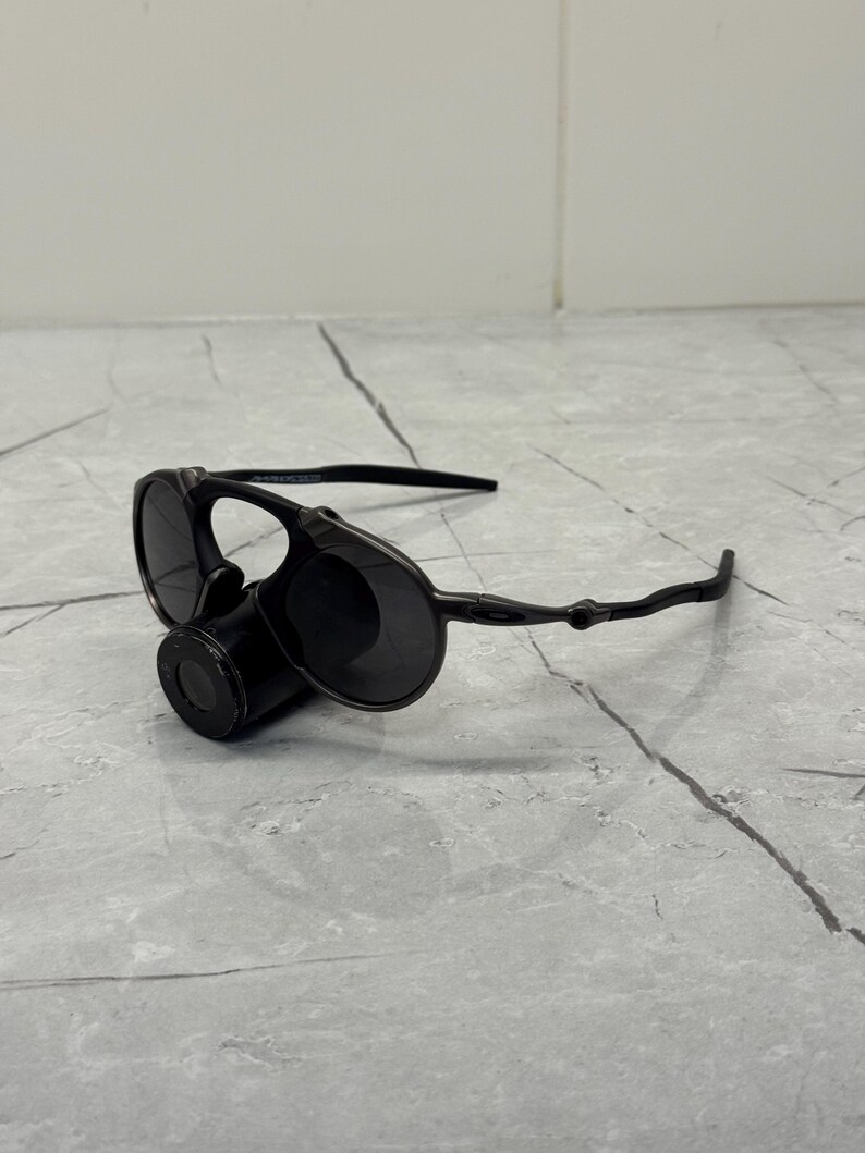 May include: Black sunglasses with a unique design. The sunglasses feature round lenses and a cylindrical component between the lenses. The arms of the sunglasses are thin and curve around the ears. The sunglasses are resting on a gray surface.
