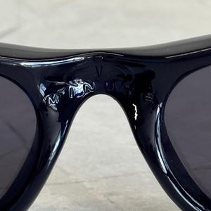 K&ouml;nnte beinhalten: Nahaufnahme einer schwarzen Sonnenbrille mit dunklen Gl&auml;sern. Die Sonnenbrille hat einen geschwungenen Rahmen, und die Buchstaben "V" und "N" sind auf dem Steg zu sehen. Die Sonnenbrille liegt auf einer hellgrauen Oberfl&auml;che.