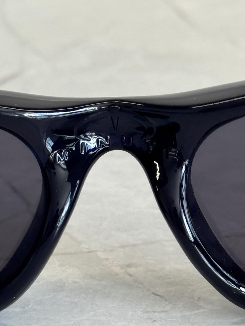 K&ouml;nnte beinhalten: Nahaufnahme einer schwarzen Sonnenbrille mit dunklen Gl&auml;sern. Die Sonnenbrille hat einen geschwungenen Rahmen, und die Buchstaben "V" und "N" sind auf dem Steg zu sehen. Die Sonnenbrille liegt auf einer hellgrauen Oberfl&auml;che.