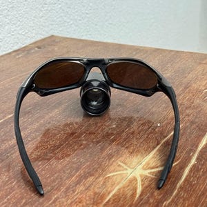 Op de afbeelding: Zwarte zonnebril met bruin getinte glazen. De zonnebril heeft een strak, modern design met een zwart montuur en zwarte pootjes. De glazen zijn ovaal van vorm en beschermen tegen de zon. De zonnebril ligt op een houten oppervlak.
