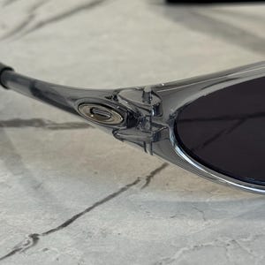 May include: Close-up of a pair of clear-framed sunglasses with dark lenses. The sunglasses have a sleek, modern design with a silver emblem on the side. The frame is transparent, revealing the intricate details of the hinge and arm.