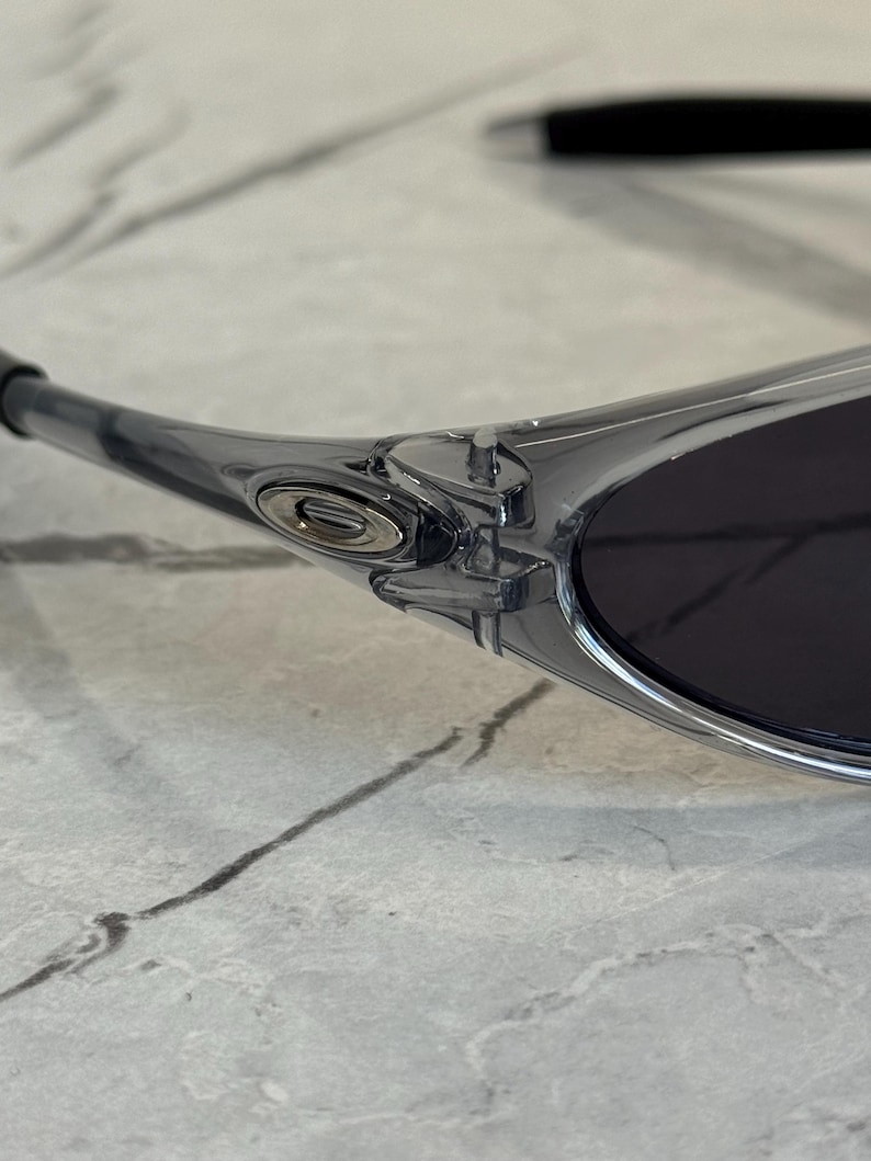 May include: Close-up of a pair of clear-framed sunglasses with dark lenses. The sunglasses have a sleek, modern design with a silver emblem on the side. The frame is transparent, revealing the intricate details of the hinge and arm.
