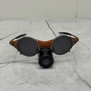 May include: Round sunglasses with dark lenses and a brown leather bridge. The frames are black with brown leather accents and stitching. The sunglasses are resting on a black cylindrical object.