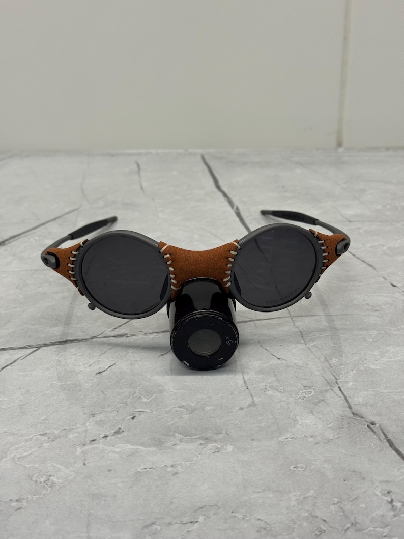 May include: Round sunglasses with dark lenses and a brown leather bridge. The frames are black with brown leather accents and stitching. The sunglasses are resting on a black cylindrical object.