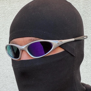 May include: Silver sunglasses with purple lenses are worn over a black balaclava. The sunglasses have a modern design. The balaclava covers the head and neck, leaving only the eyes visible. The sunglasses are a stylish accessory.