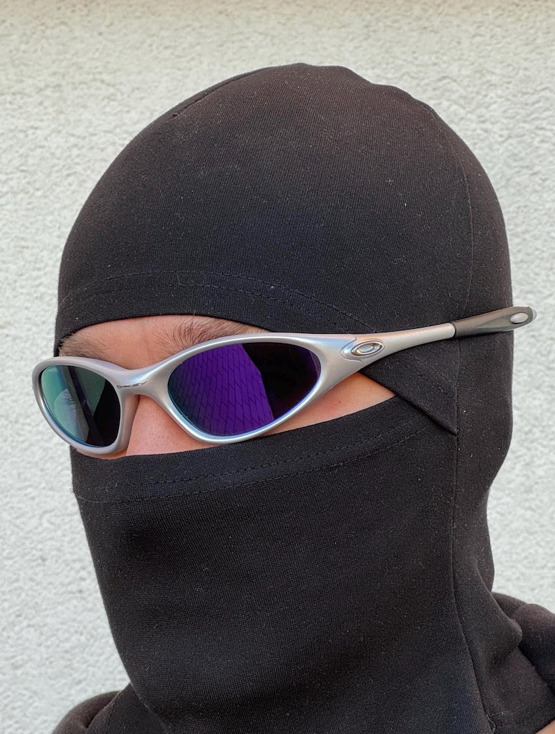 May include: Silver sunglasses with purple lenses are worn over a black balaclava. The sunglasses have a modern design. The balaclava covers the head and neck, leaving only the eyes visible. The sunglasses are a stylish accessory.