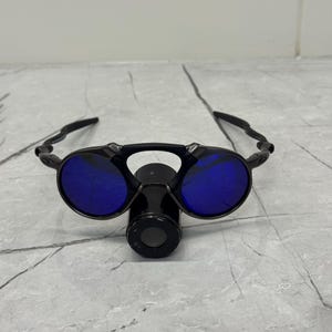 以下が含まれることがあります： 丸い青いレンズとユニークなデザインの黒いサングラス。 サングラスは、中央の円筒形のコンポーネントと湾曲したアームを備えています。 レンズは鮮やかな青色で、フレームは黒色です。