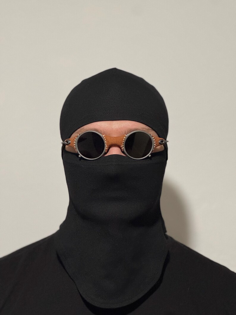 May include: A person wearing a black balaclava and goggles. The goggles have round, dark lenses, a brown leather bridge, and silver-colored metal detailing. The balaclava covers the head and neck.