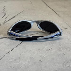 Puede incluir: Gafas de sol plateadas con lentes oscuras. Las gafas tienen un dise&ntilde;o deportivo con detalles azules en las patillas y un puente nasal negro. Las gafas se muestran sobre una superficie gris claro.