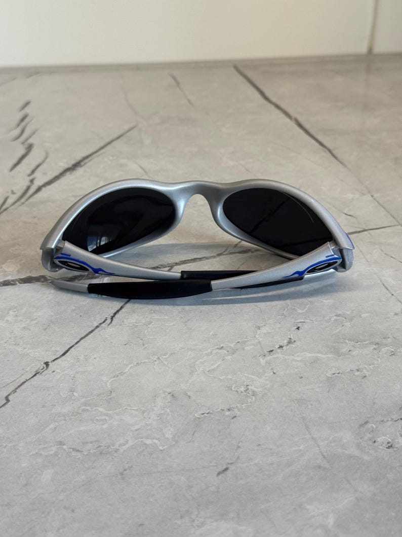 Puede incluir: Gafas de sol plateadas con lentes oscuras. Las gafas tienen un dise&ntilde;o deportivo con detalles azules en las patillas y un puente nasal negro. Las gafas se muestran sobre una superficie gris claro.