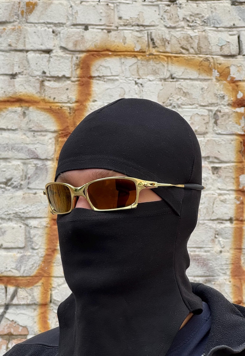 May include: Gold-framed sunglasses with brown lenses. The sunglasses are worn with a black balaclava. The background is a weathered brick wall with patches of white and orange.