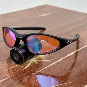 May include: Black sunglasses with orange-tinted lenses. The sunglasses have a sleek, curved design with a small logo on the temple. The lenses reflect a mix of colors, and the frame is a matte black.