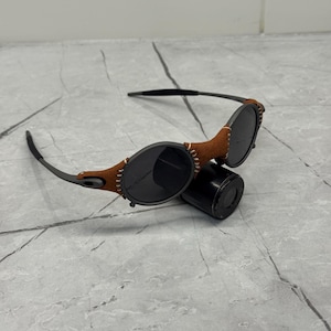 May include: Black sunglasses with round lenses and a brown leather accent stitched onto the frame. The sunglasses have a sleek, modern design and are resting on a black cylindrical object.
