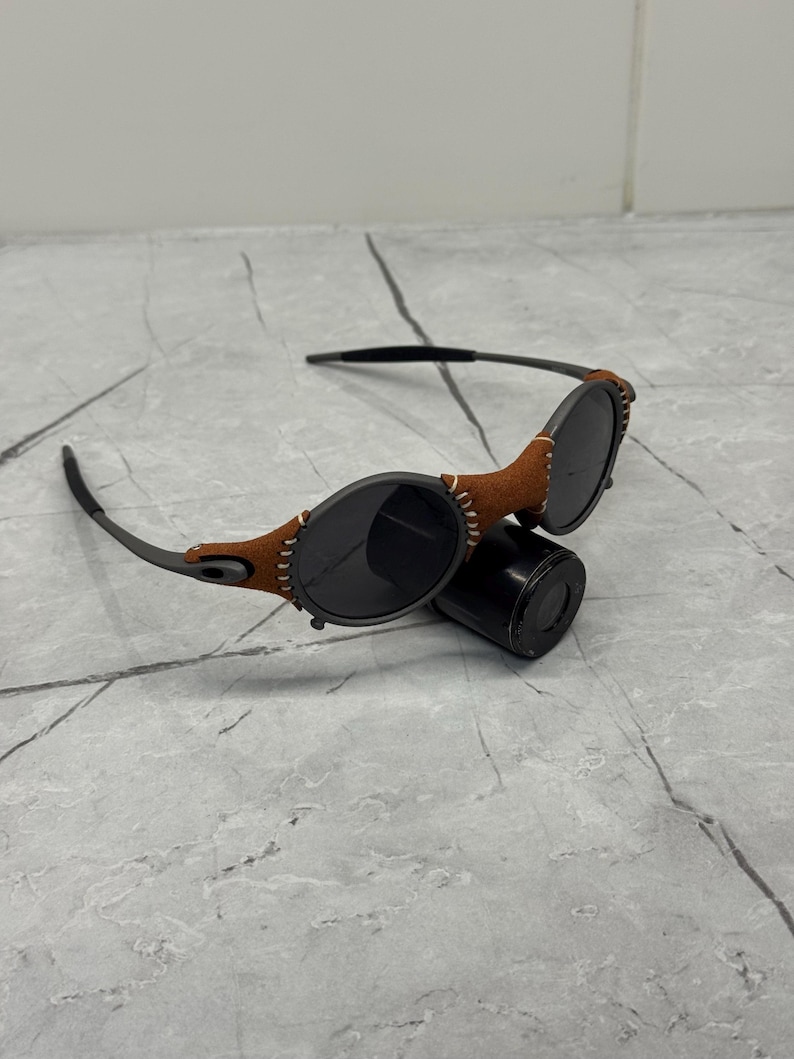 May include: Black sunglasses with round lenses and a brown leather accent stitched onto the frame. The sunglasses have a sleek, modern design and are resting on a black cylindrical object.