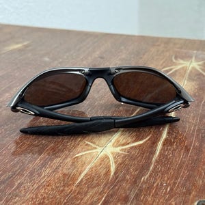 Op de afbeelding: Zwarte zonnebril met bruine glazen, opengeklapt op een houten oppervlak. De zonnebril heeft een strak, modern design met een zwart montuur en donkere glazen. De pootjes zijn opgevouwen, waardoor het scharnier en de details zichtbaar zijn.