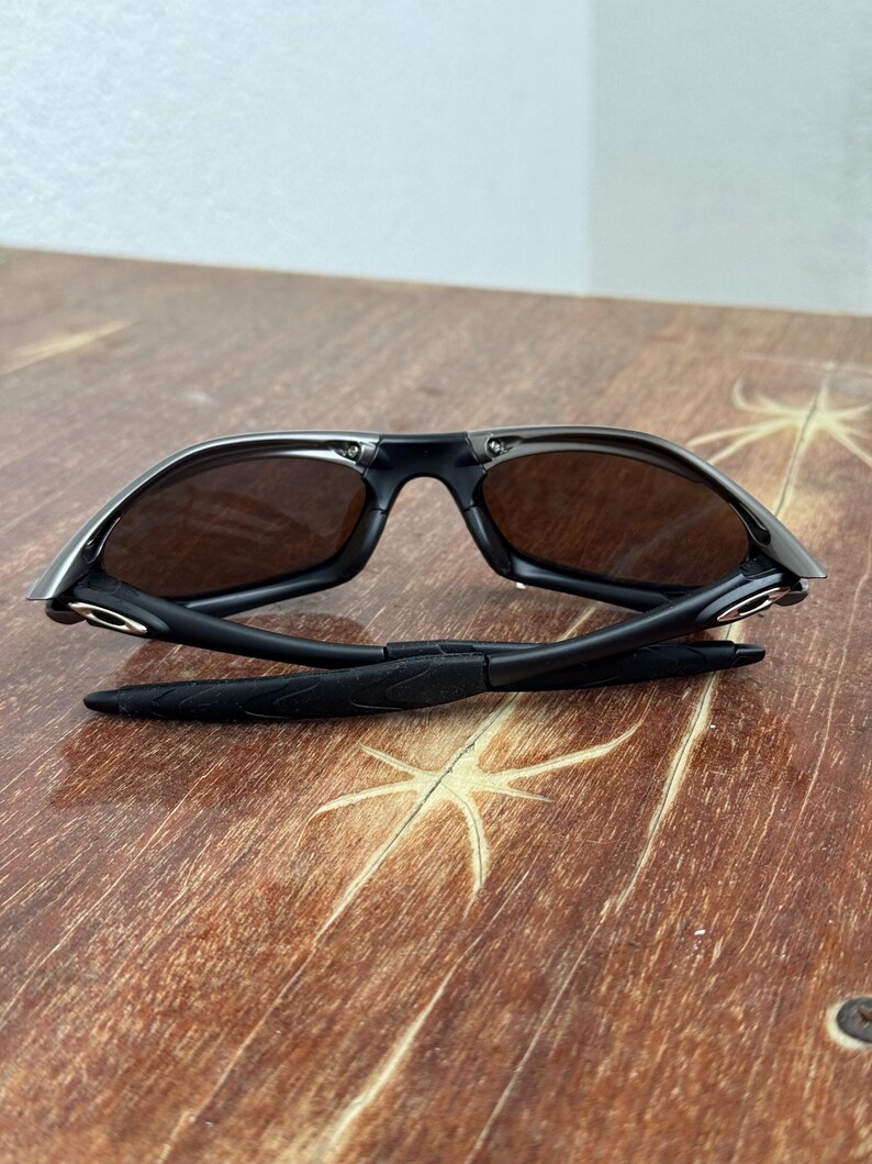 Op de afbeelding: Zwarte zonnebril met bruine glazen, opengeklapt op een houten oppervlak. De zonnebril heeft een strak, modern design met een zwart montuur en donkere glazen. De pootjes zijn opgevouwen, waardoor het scharnier en de details zichtbaar zijn.
