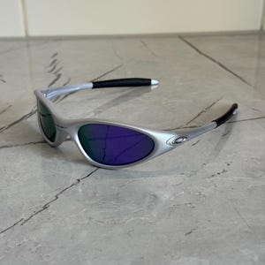May include: Silver sunglasses with purple lenses. The sunglasses have a streamlined design, with black detailing on the arms. The lenses reflect a green colour in some lights, adding a distinctive element to the eyewear.