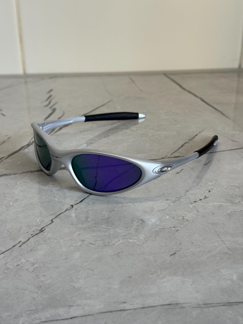 May include: Silver sunglasses with purple lenses. The sunglasses have a streamlined design, with black detailing on the arms. The lenses reflect a green colour in some lights, adding a distinctive element to the eyewear.