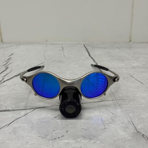 Może przedstawiać: Okulary przeciwsłoneczne w srebrnej oprawie z okrągłymi, niebieskimi soczewkami. Okulary mają unikalny design z czarnym mostkiem nosowym i zausznikami. Soczewki odbijają jasnoniebieski kolor, a oprawka jest metaliczna srebrna.