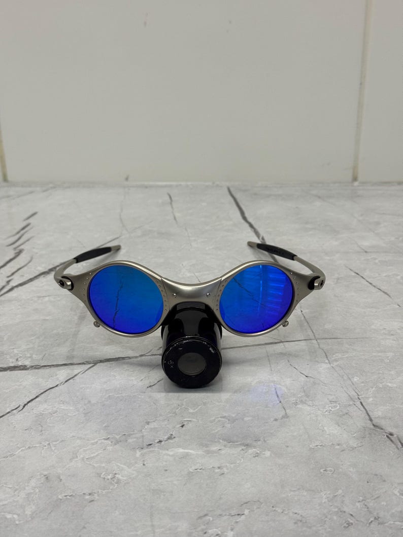 Może przedstawiać: Okulary przeciwsłoneczne w srebrnej oprawie z okrągłymi, niebieskimi soczewkami. Okulary mają unikalny design z czarnym mostkiem nosowym i zausznikami. Soczewki odbijają jasnoniebieski kolor, a oprawka jest metaliczna srebrna.