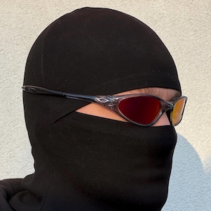 K&ouml;nnte beinhalten: Eine Person tr&auml;gt eine schwarze Sturmhaube und eine Sonnenbrille mit rot get&ouml;nten Gl&auml;sern. Die Sonnenbrille hat einen transparenten Rahmen und dunkle B&uuml;gel. Die Sturmhaube bedeckt Kopf und Hals, wobei nur die Augen sichtbar sind.