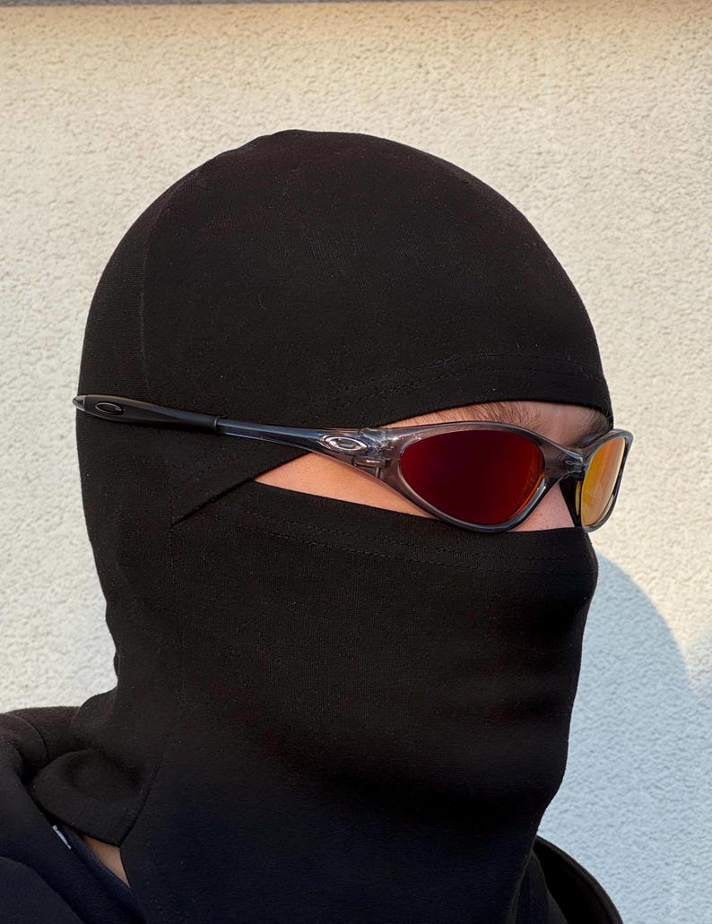 K&ouml;nnte beinhalten: Eine Person tr&auml;gt eine schwarze Sturmhaube und eine Sonnenbrille mit rot get&ouml;nten Gl&auml;sern. Die Sonnenbrille hat einen transparenten Rahmen und dunkle B&uuml;gel. Die Sturmhaube bedeckt Kopf und Hals, wobei nur die Augen sichtbar sind.