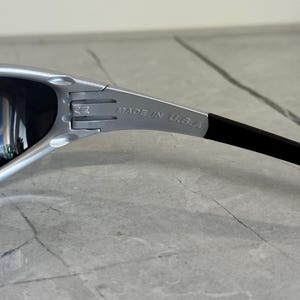 May include: Silver-framed sunglasses with dark lenses. The temple arm is black and features the text "MADE IN U.S.A." The sunglasses have a sporty, modern design.