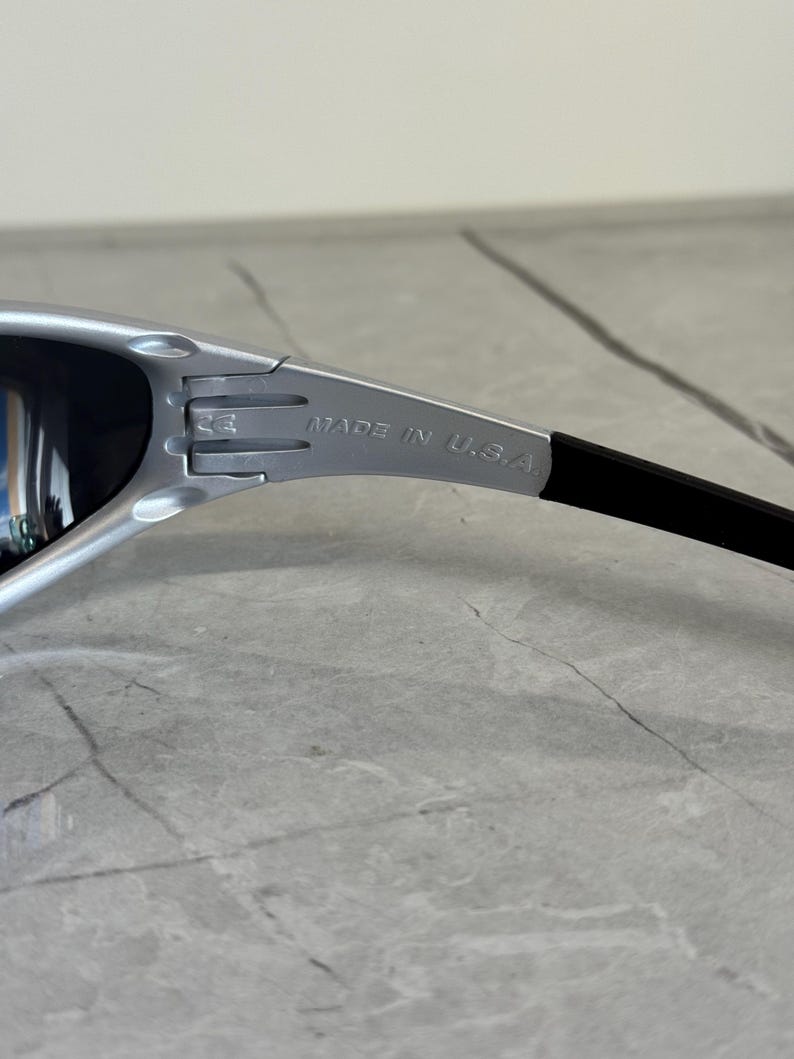 May include: Silver-framed sunglasses with dark lenses. The temple arm is black and features the text "MADE IN U.S.A." The sunglasses have a sporty, modern design.