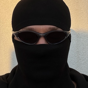 May include: A person wearing a black balaclava and dark sunglasses. The balaclava covers the head and neck, with openings for the eyes. The sunglasses have a clear frame and dark lenses. The background is a light beige color.