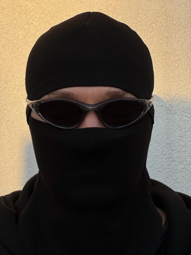 May include: A person wearing a black balaclava and dark sunglasses. The balaclava covers the head and neck, with openings for the eyes. The sunglasses have a clear frame and dark lenses. The background is a light beige color.