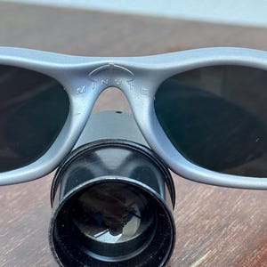 May include: Close-up of gray sunglasses with dark lenses and the word "MINUTE" printed on the frame. A black cylindrical object is positioned below the glasses. The image is taken on a wooden surface.