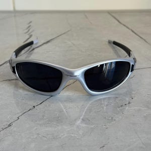 May include: Silver-framed sunglasses with black lenses. The sunglasses have a sleek, sporty design with black arms and a curved shape. The brand name "Oakley" is visible on the frame. The sunglasses are designed to protect the eyes from sunlight.