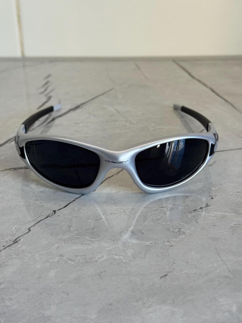 May include: Silver-framed sunglasses with black lenses. The sunglasses have a sleek, sporty design with black arms and a curved shape. The brand name "Oakley" is visible on the frame. The sunglasses are designed to protect the eyes from sunlight.