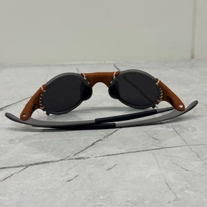 May include: Sunglasses with dark lenses and a brown leather bridge. The arms are gray with a black accent. The leather bridge is stitched with white thread. The sunglasses are on a gray surface.