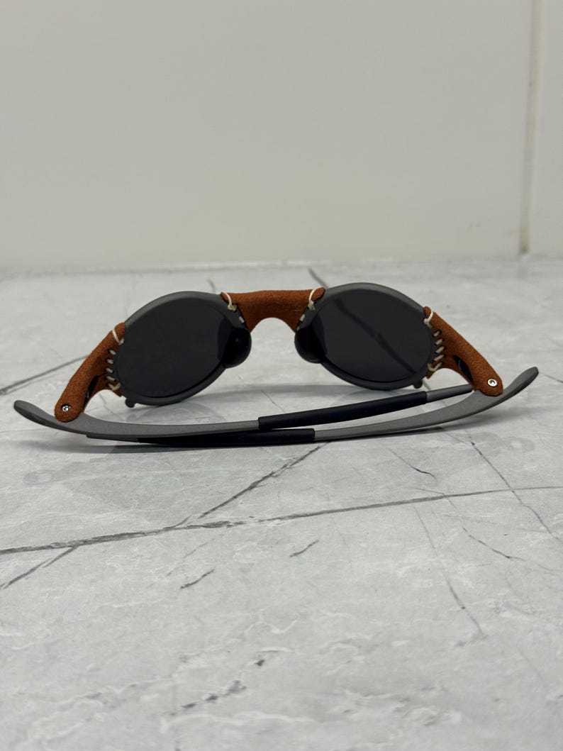 May include: Sunglasses with dark lenses and a brown leather bridge. The arms are gray with a black accent. The leather bridge is stitched with white thread. The sunglasses are on a gray surface.