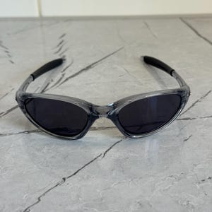 May include: Clear-framed sunglasses with dark lenses. The sunglasses have a sleek, sporty design with curved temples and a wraparound style. The lenses are a dark, solid color, providing shade and protection from the sun.