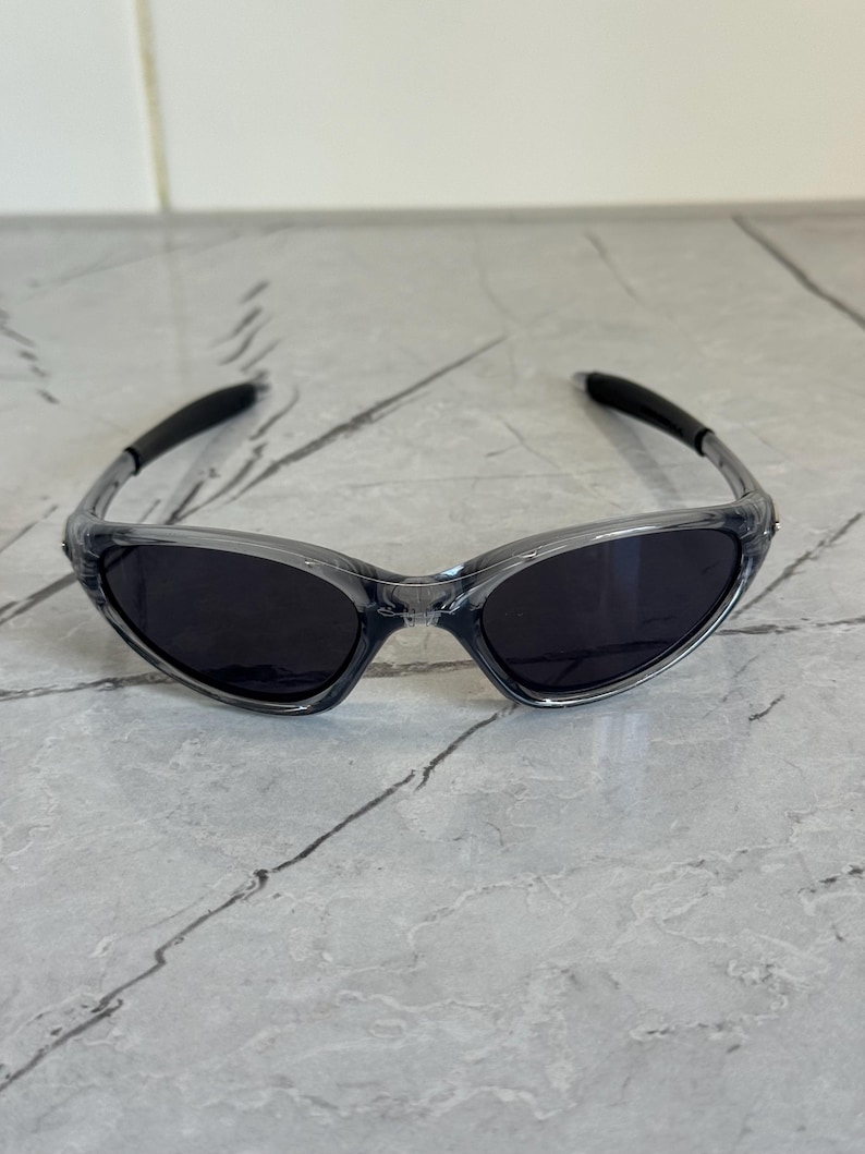 May include: Clear-framed sunglasses with dark lenses. The sunglasses have a sleek, sporty design with curved temples and a wraparound style. The lenses are a dark, solid color, providing shade and protection from the sun.