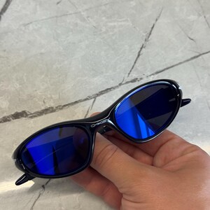 May include: Black sunglasses with blue lenses. The sunglasses have a sleek, oval shape and a glossy finish. The lenses reflect a bright blue color. The frame has the brand name printed on the side.