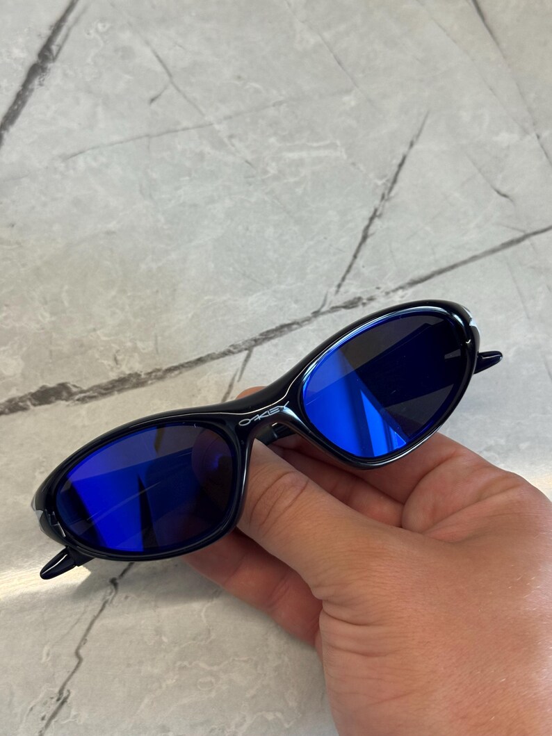 May include: Black sunglasses with blue lenses. The sunglasses have a sleek, oval shape and a glossy finish. The lenses reflect a bright blue color. The frame has the brand name printed on the side.