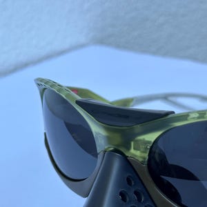 May include: Close-up of olive green sunglasses with dark lenses. The sunglasses have a sporty design with a black nose bridge and side shields. The frame is a translucent green color.