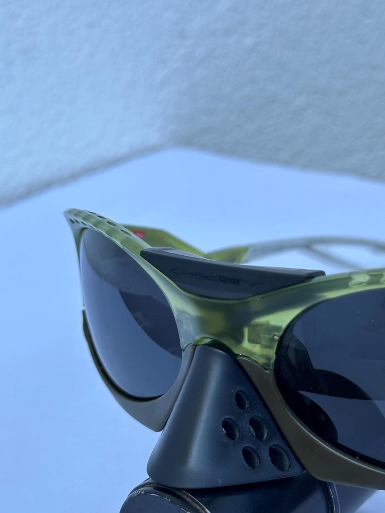 May include: Close-up of olive green sunglasses with dark lenses. The sunglasses have a sporty design with a black nose bridge and side shields. The frame is a translucent green color.