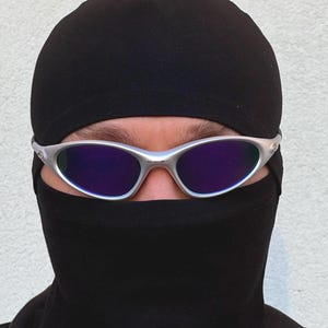 May include: Silver-framed sunglasses with purple lenses are worn over a black balaclava. The balaclava covers the head and neck, with only the eyes and eyebrows visible. The background is a plain, light-coloured wall.