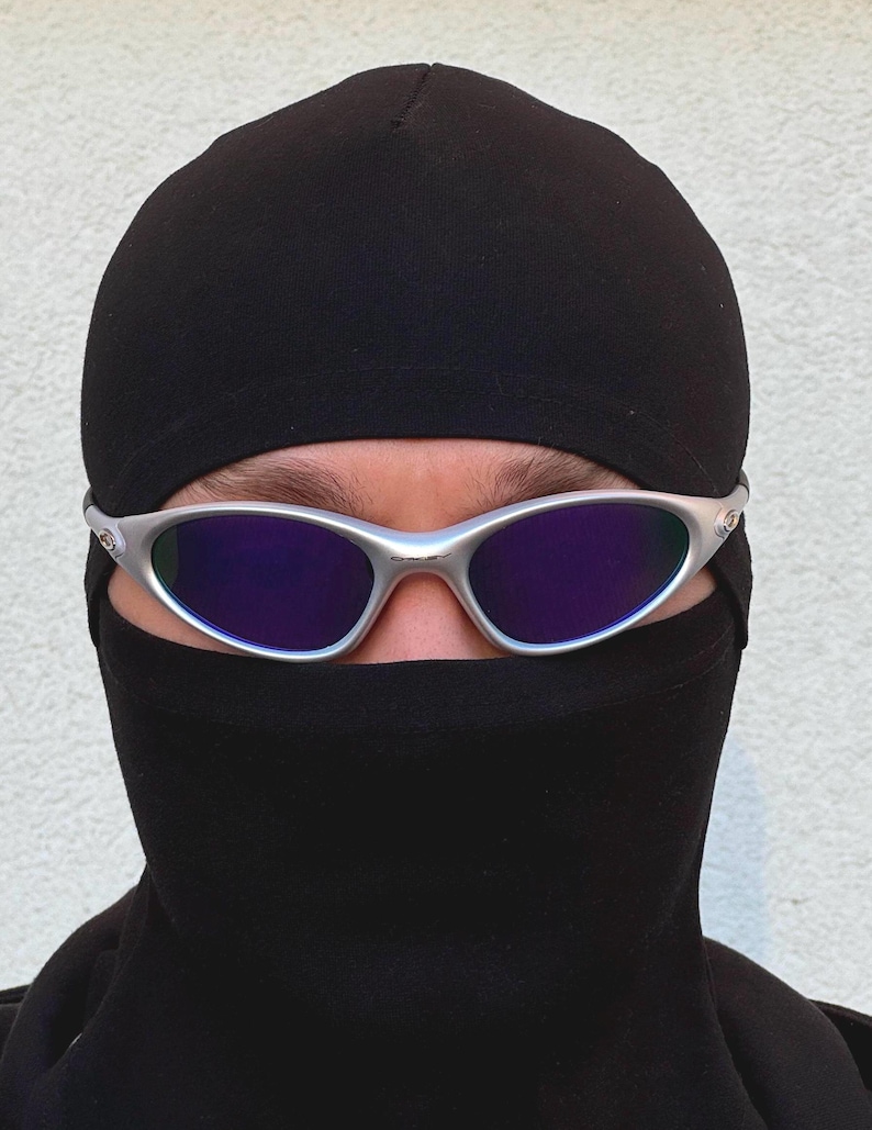 May include: Silver-framed sunglasses with purple lenses are worn over a black balaclava. The balaclava covers the head and neck, with only the eyes and eyebrows visible. The background is a plain, light-coloured wall.