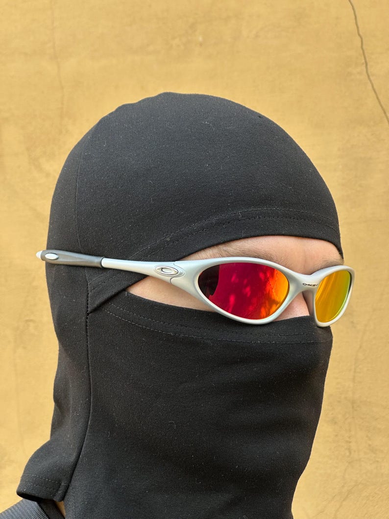 May include: Silver-framed sunglasses with red and orange lenses are worn over a black balaclava. The sunglasses have a sleek, modern design. The balaclava covers the head and neck, leaving only the eyes visible.