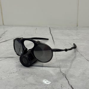 May include: Black sunglasses with a distinctive design. The sunglasses feature round lenses and a unique bridge with a cylindrical component. The arms are thin and curve around the ears. The lenses are dark, providing shade.