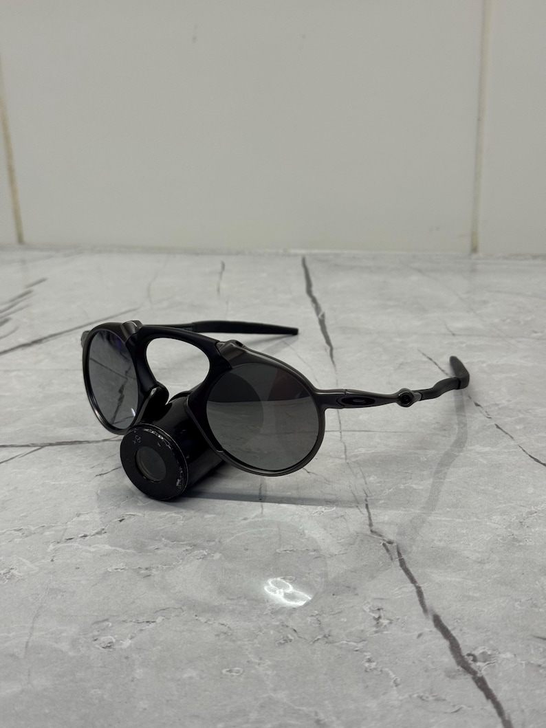 May include: Black sunglasses with a distinctive design. The sunglasses feature round lenses and a unique bridge with a cylindrical component. The arms are thin and curve around the ears. The lenses are dark, providing shade.
